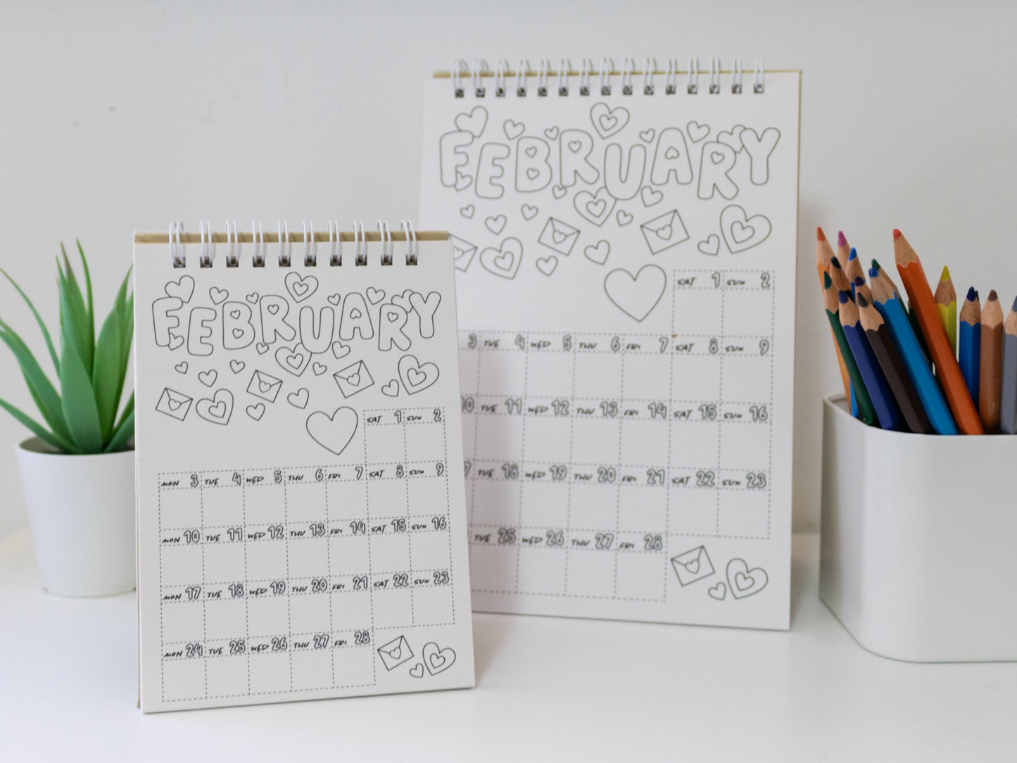 Two 2026 colouring desk calendar design on a white surface with coloured pencils - February pages