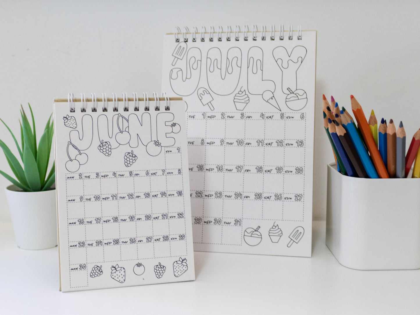 Two 2026 colouring desk calendars on a white surface with coloured pencils - June and July pages