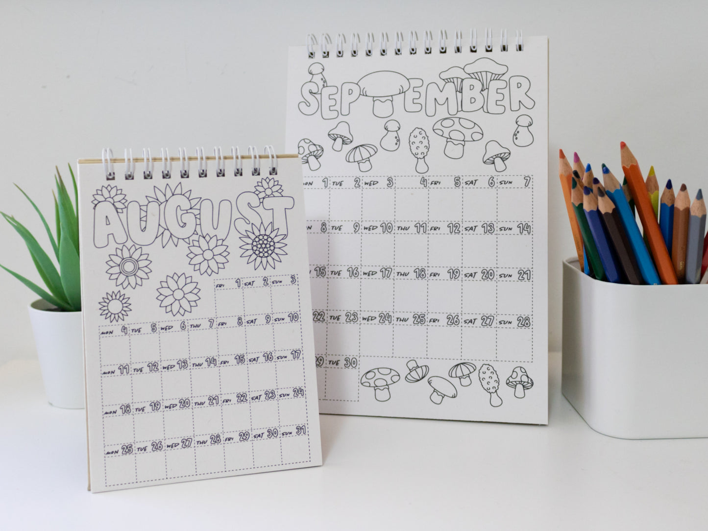 Two 2026 colouring desk calendars on a white surface with coloured pencils - August & September pages