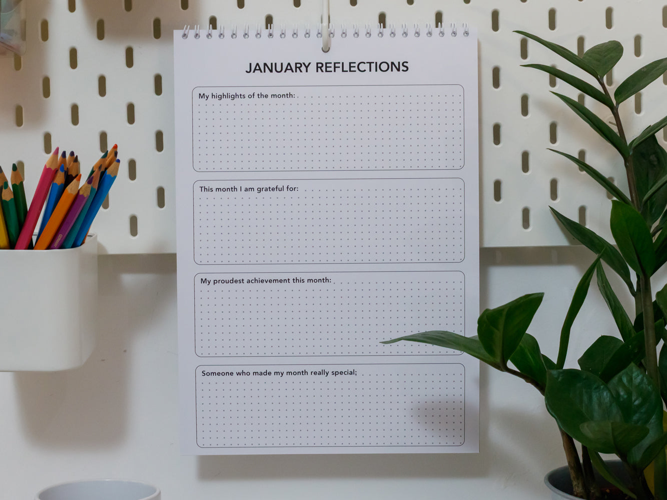 2026 Wall Calendar: January reflections page on a white pegboard with stationery items and a plant.
