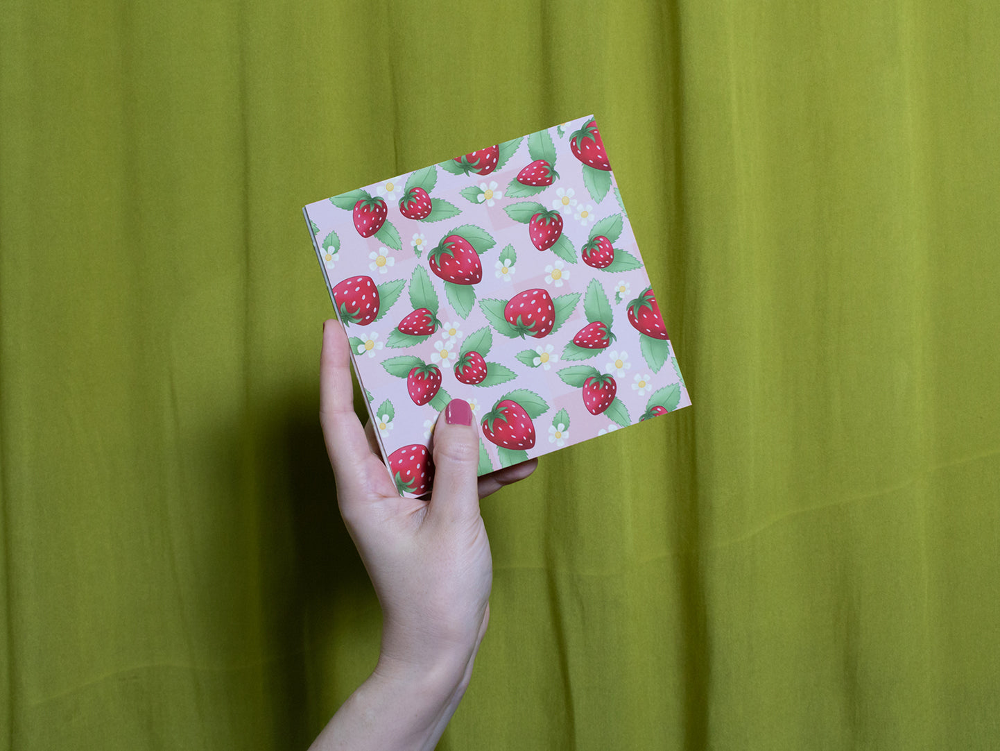 Hand holding a sketchbook with strawberry pattern against a green curtain