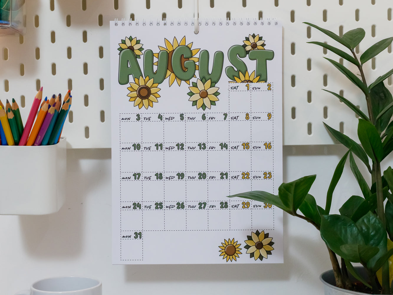 2026 Wall Calendar: August page and sunflower designs on a white wall with stationery items.