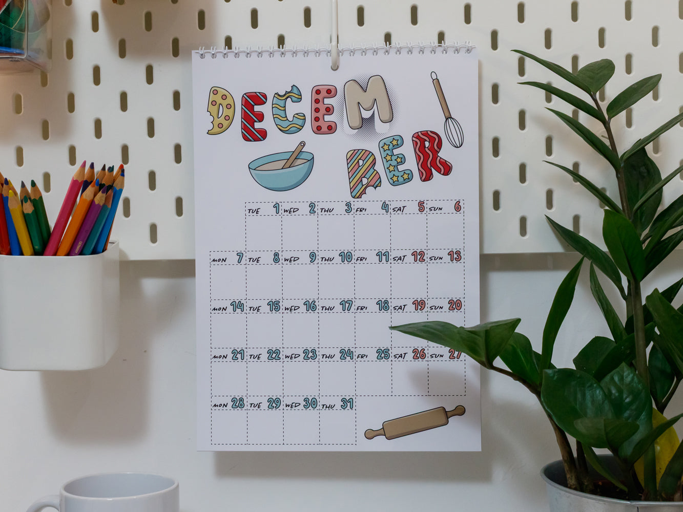 2026 Wall Calendar: December page on a pegboard with stationery items and a plant in the background