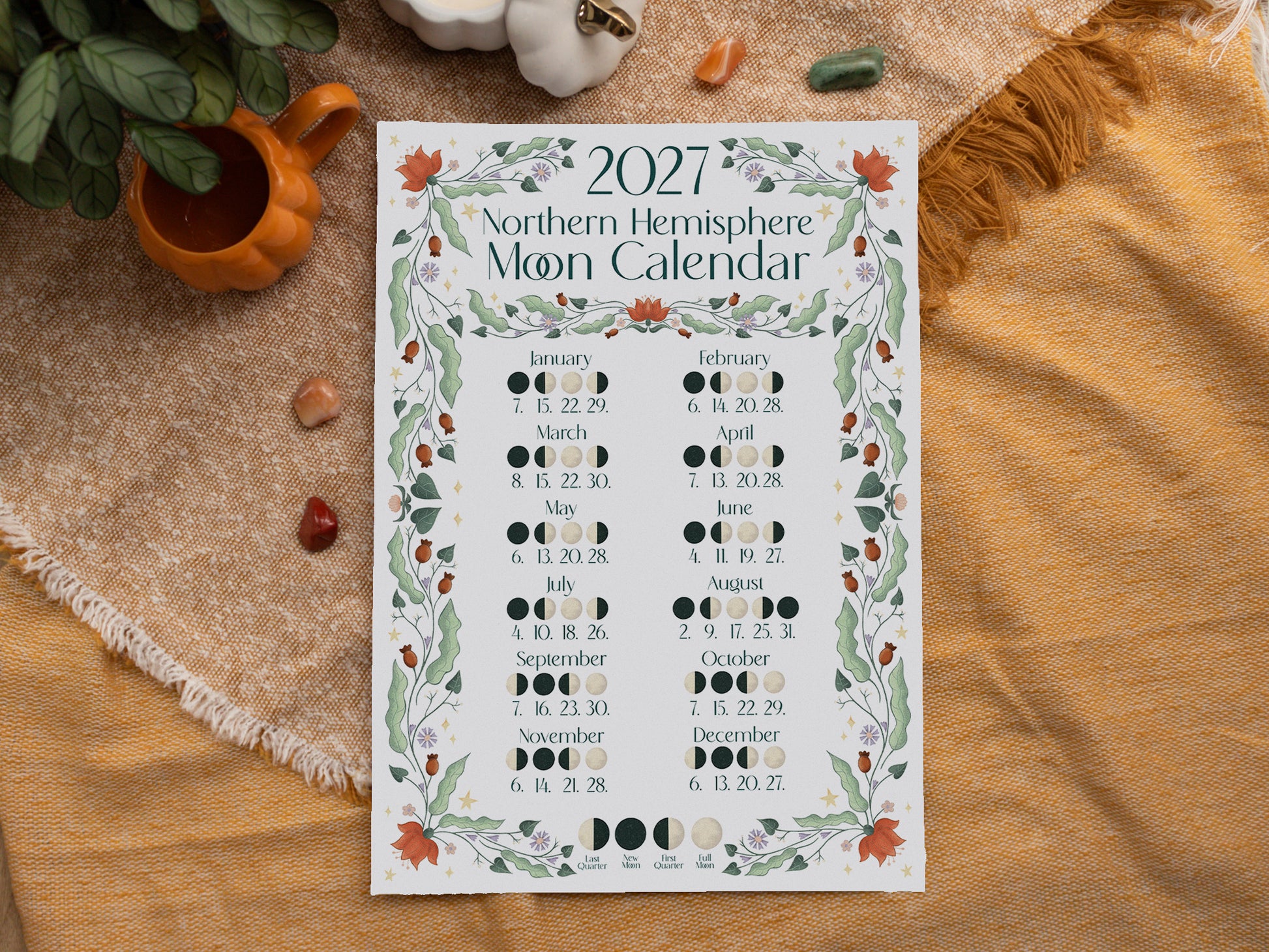 A 2027 Northern Hemisphere Moon Calendar print laid flat on a textured mustard-yellow blanket with autumn styling props including a small orange pot, green leaves, and a white pumpkin.