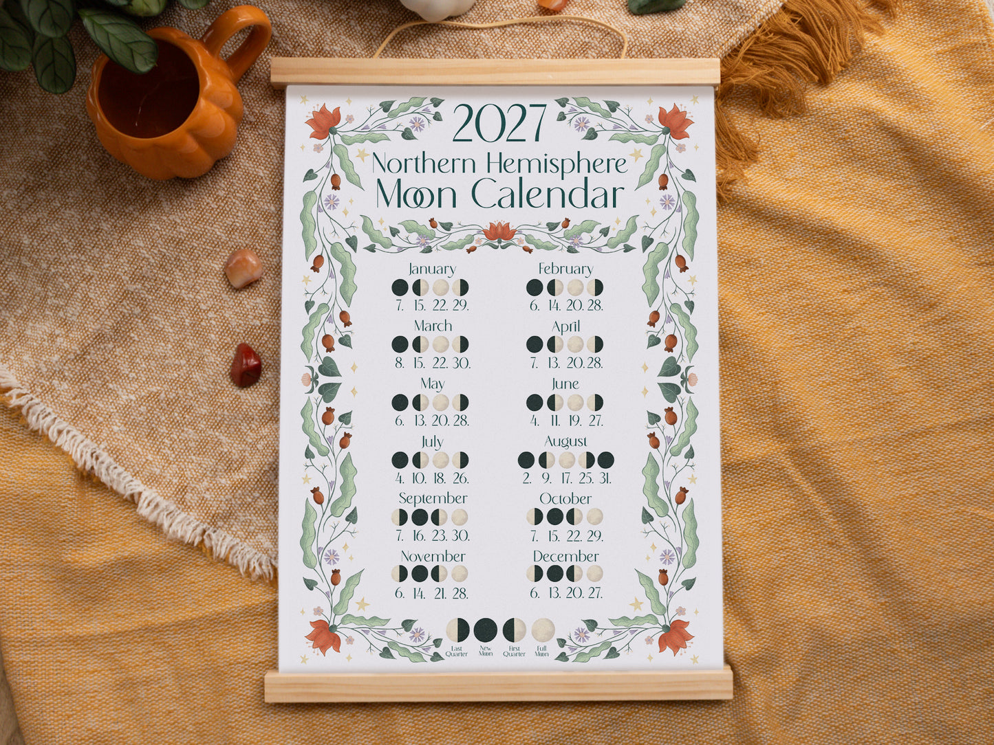 A 2027 Northern Hemisphere Moon Calendar hanging on a rustic burlap surface with autumn props. The calendar features a white background with a decorative border of green leaves and orange maple leaves, displaying the moon phases for each day of the year.