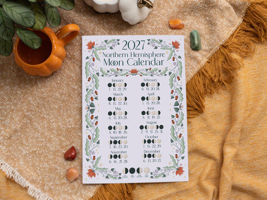 A 2027 Northern Hemisphere Moon Calendar print is displayed on a textured beige and mustard yellow blanket with autumn-themed props including a small orange pumpkin, crystals, and a white ceramic ornament.