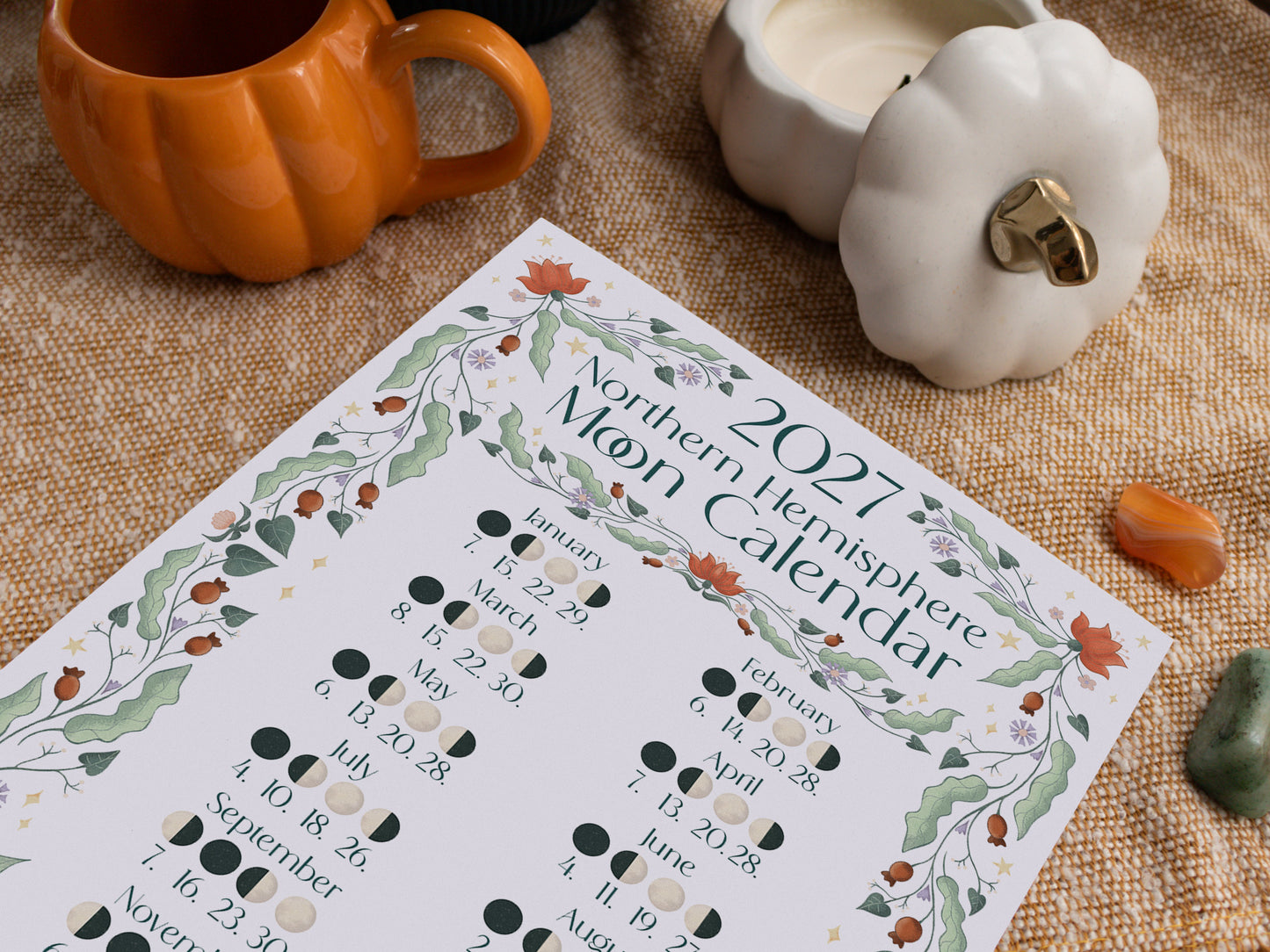 A 2027 Northern Hemisphere Moon Calendar sheet laid on a woven beige fabric, surrounded by autumn-themed props including an orange pumpkin-shaped mug, a white pumpkin candle holder, and small crystals.
