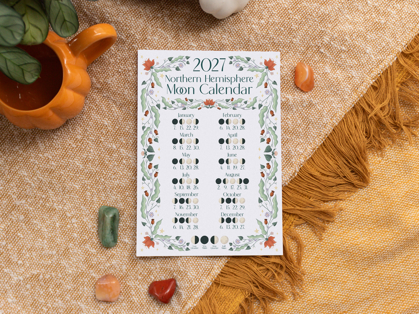 A 2027 Moon Calendar print is displayed in a styled flat-lay setting on a textured beige fabric, surrounded by crystals and a small orange mug.