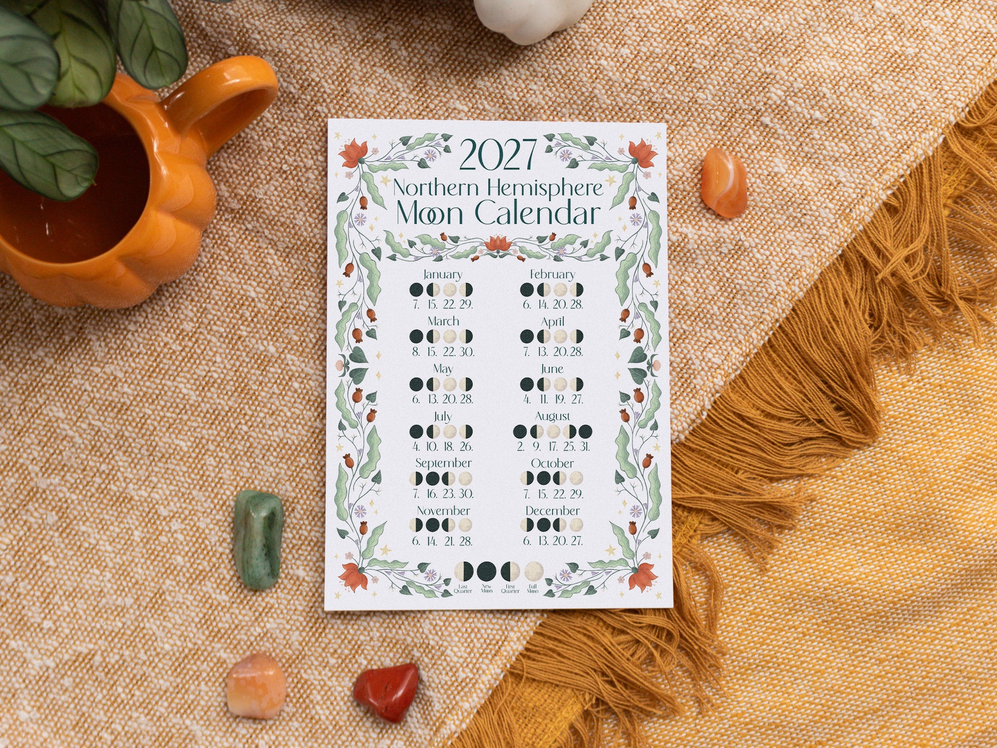 A 2027 Moon Calendar print is displayed in a styled flat-lay setting on a textured beige fabric, surrounded by crystals and a small orange mug.