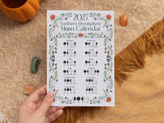 A hand holds a 2027 Moon Calendar print, which features a floral border and moon phase charts. The print is styled on a textured beige fabric with autumn props like a small pumpkin and crystals.