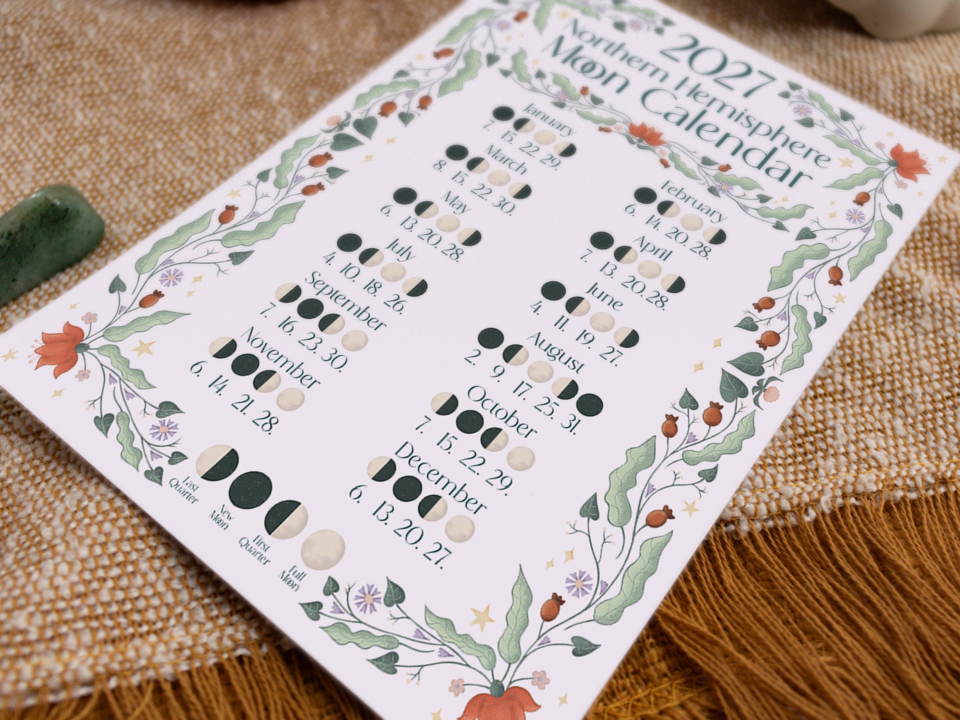 A printed Moon Calendar for 2027 lies on a textured fabric surface. The white card features a decorative border of illustrated green leaves, orange flowers, and stars, with columns of moon phase diagrams and dates for each month.
