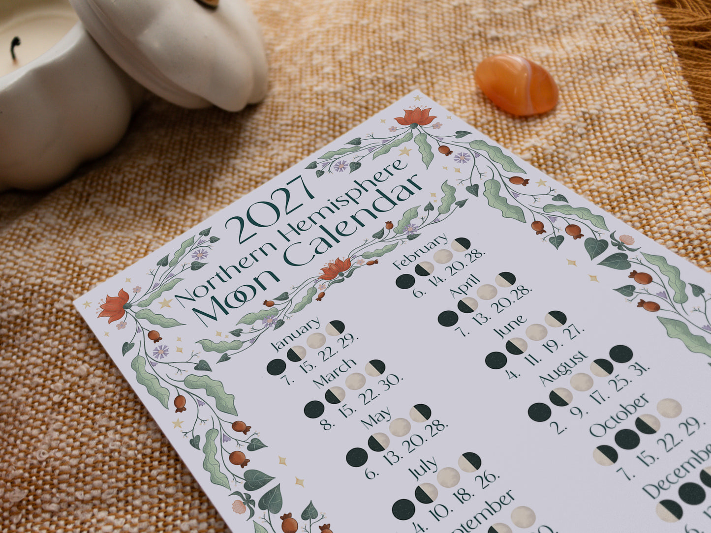 A 2027 Northern Hemisphere Moon Calendar print laid on a woven beige fabric surface, with a candle, small white pumpkin, and amber crystal nearby.