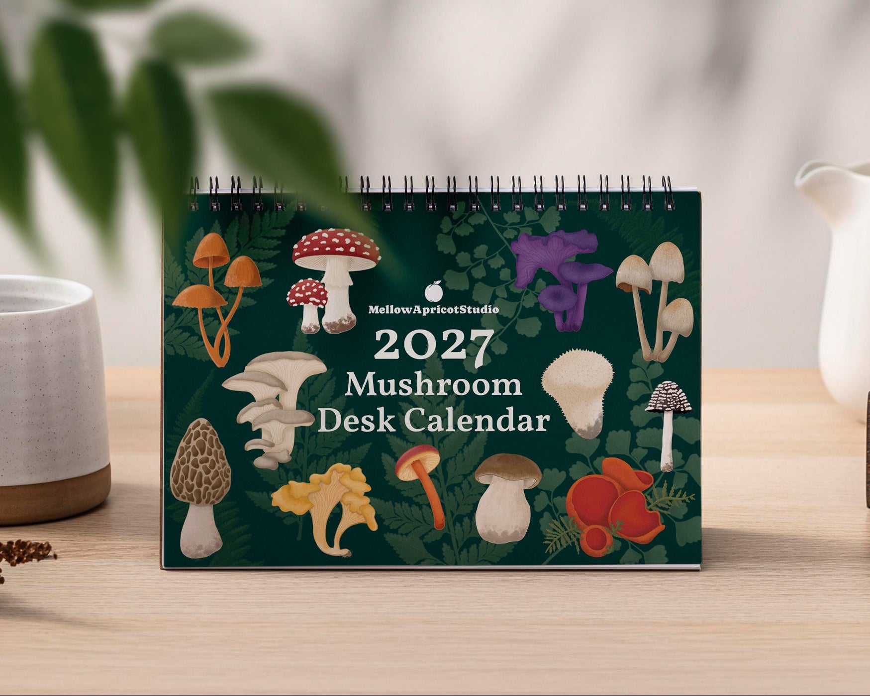 A 2027 Mushroom Desk Calendar is displayed on a light wood tabletop. The calendar cover is dark green with various colourful illustrated mushrooms and features a spiral binding at the top.