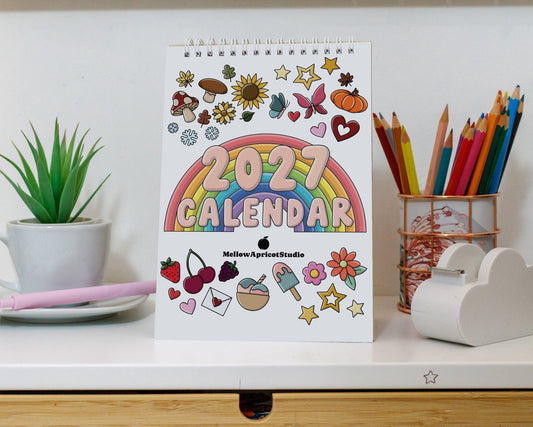 A 2027 desk calendar with colorful cartoon illustrations stands on a white desk. The calendar features a rainbow design in the center and is surrounded by decorative stickers.