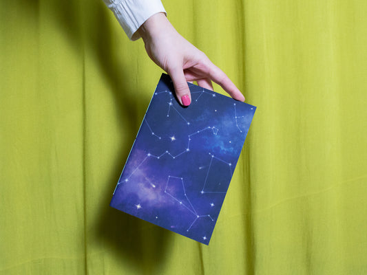 Hand holding a blue sketchbook with star constellation design against a green curtain background