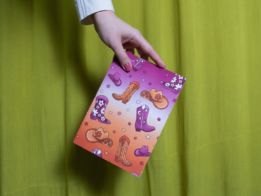 Hand holding a colourful sketchbook with cowboy boot designs against a green curtain background