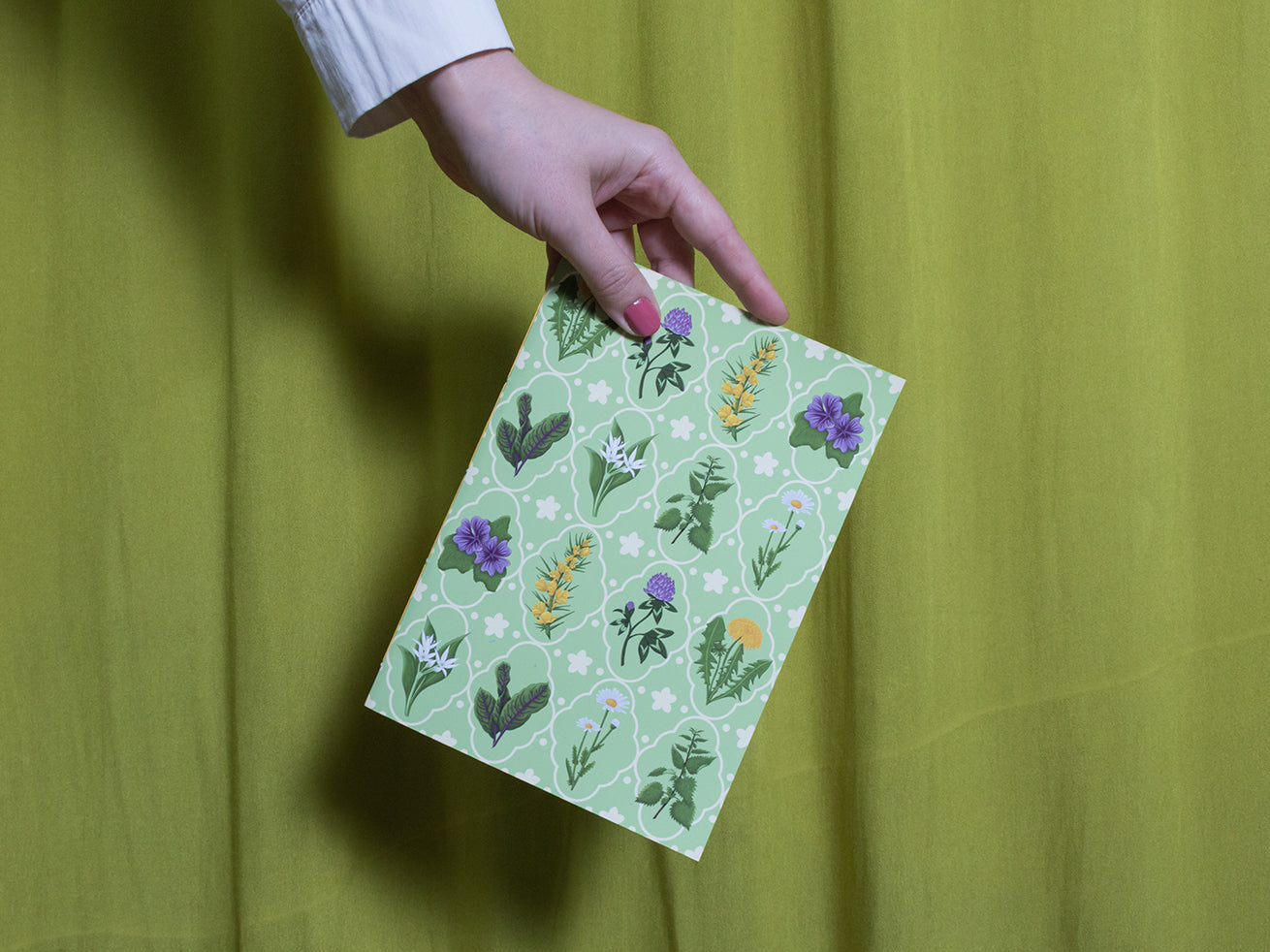 Hand holding a green sketchbook with floral patterns against a green background
