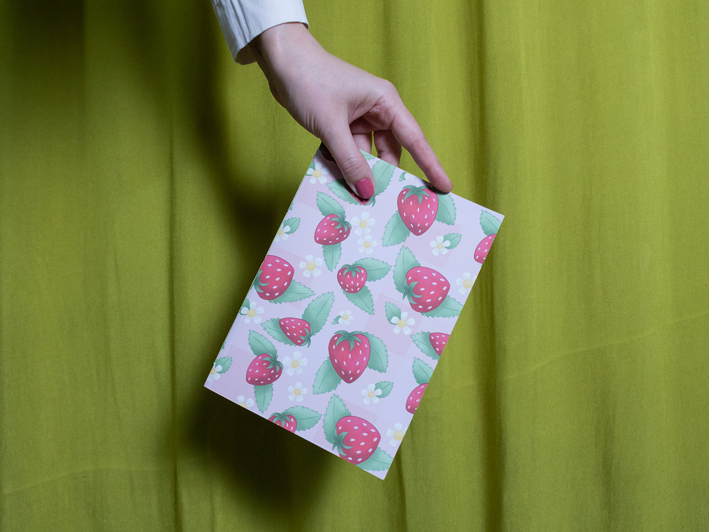 Hand holding a pink sketchbook with strawberry pattern against a green curtain background