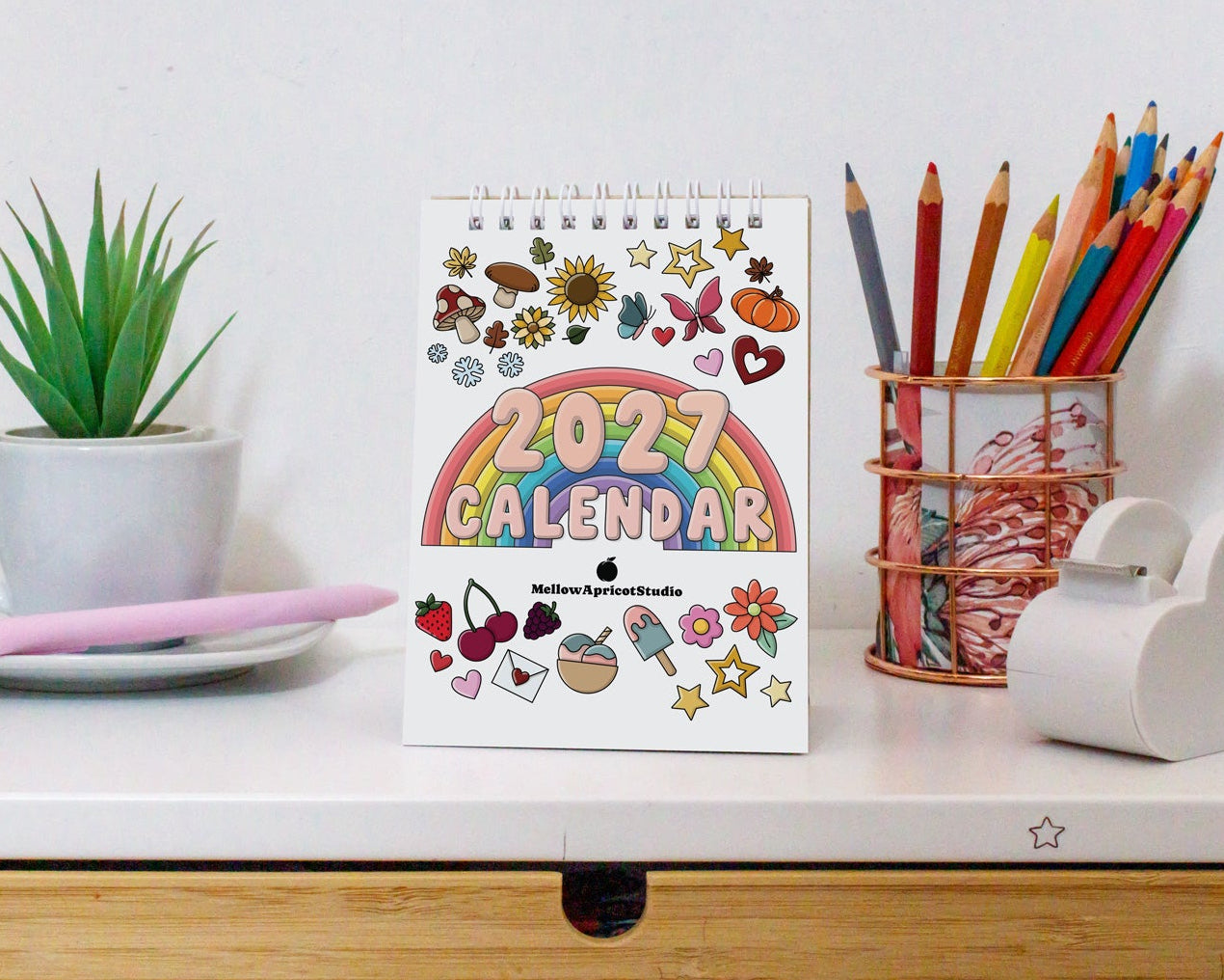 A 2027 spiral-bound planner calendar with colorful illustrations and a rainbow design, styled on a white desk next to a small potted plant, a pink pen, and a pencil cup.