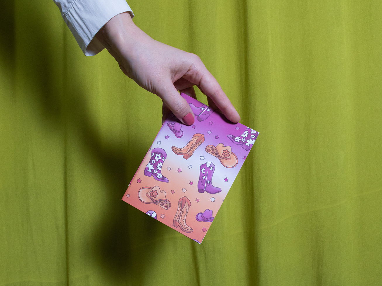 Hand holding a colourful sketchbook with howdy design against a green curtain background