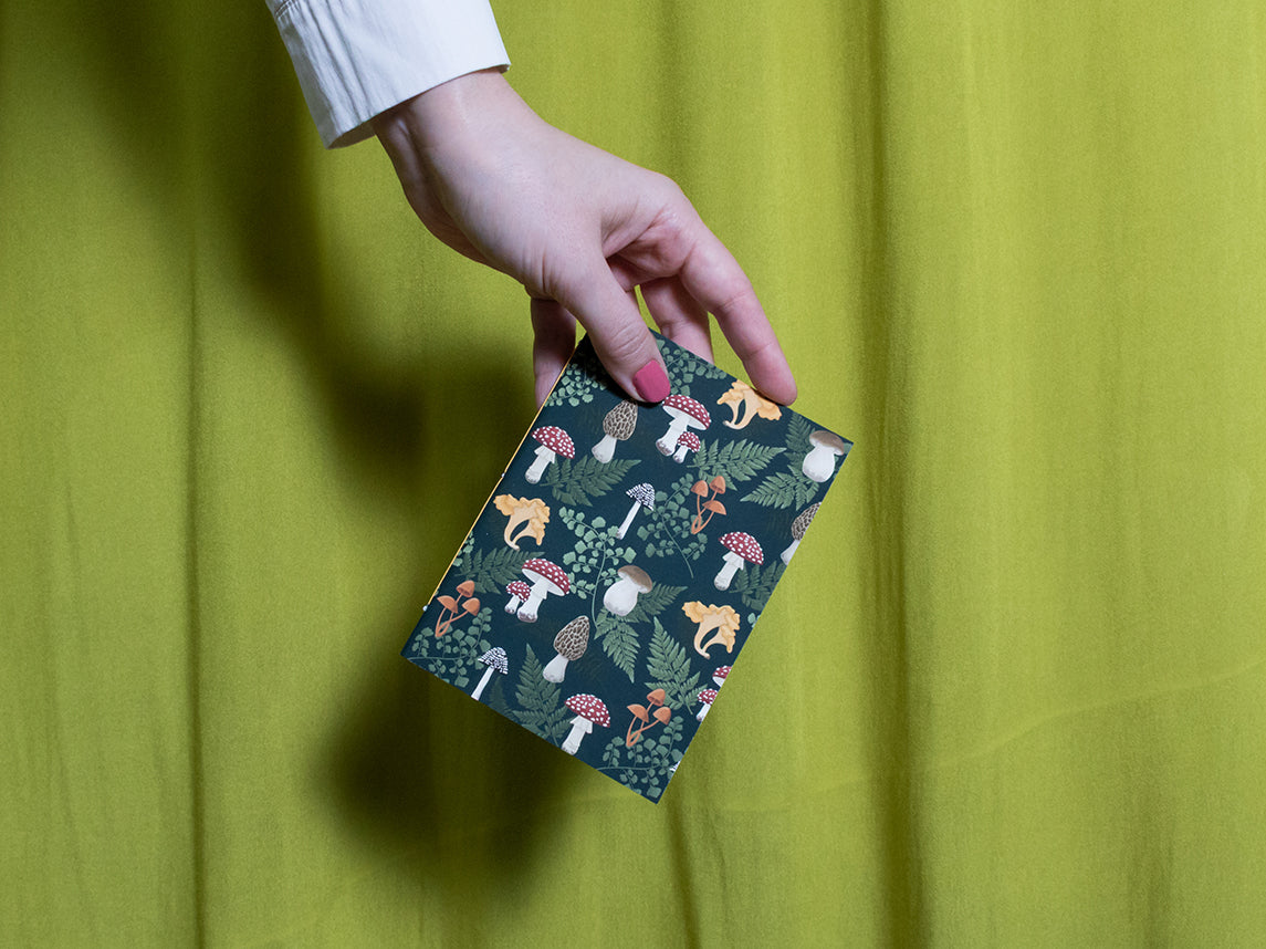 Hand holding a small sketchbook with mushroom design against a green background