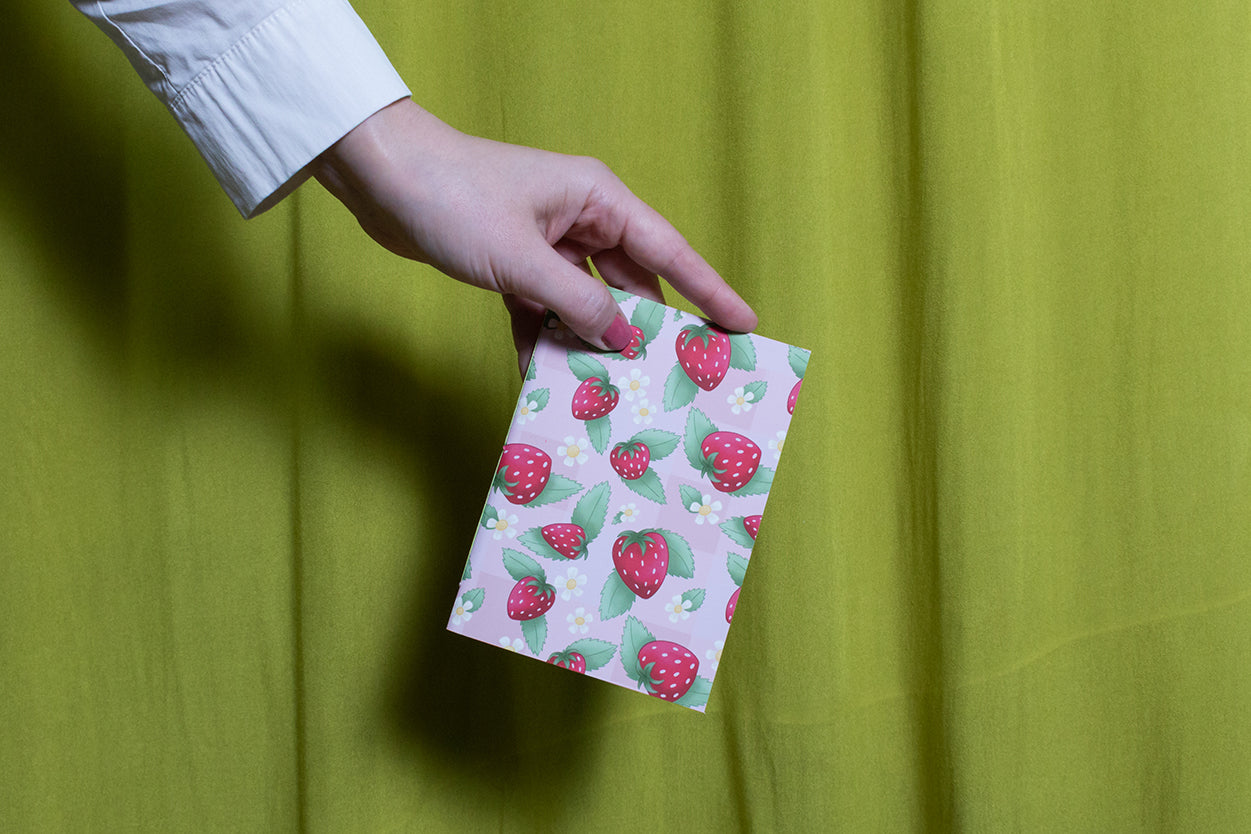 Hand holding a small sketchbook with strawberry pattern against a green curtain background