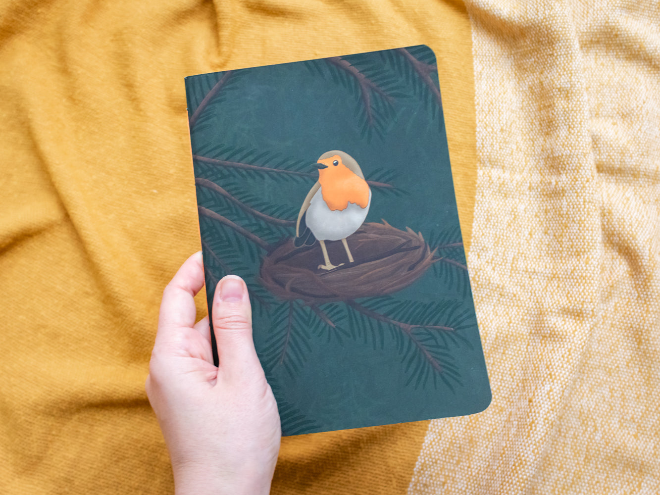 Hand holding a notebook with a robin illustration on a textured yellow fabric background