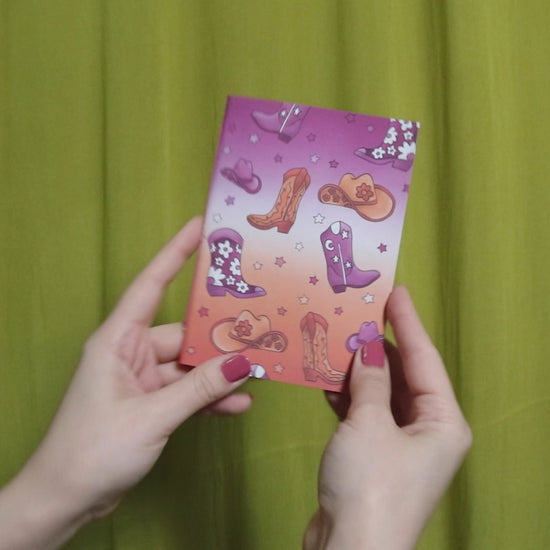 Video of two hands holding and flipping through a small sketchbook with howdy pattern against a green curtain background