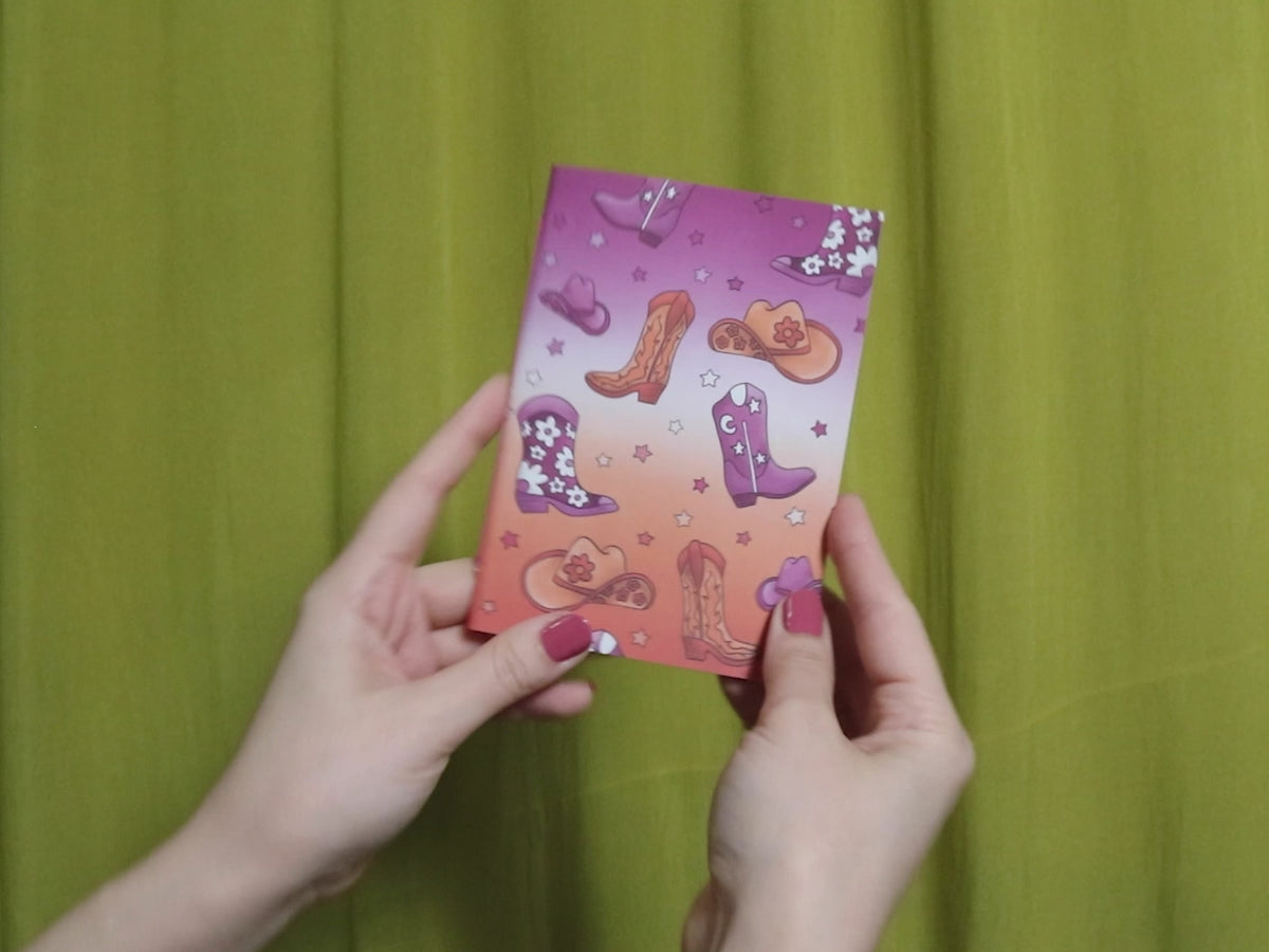 Video of two hands holding and flipping through a small sketchbook with howdy pattern against a green curtain background