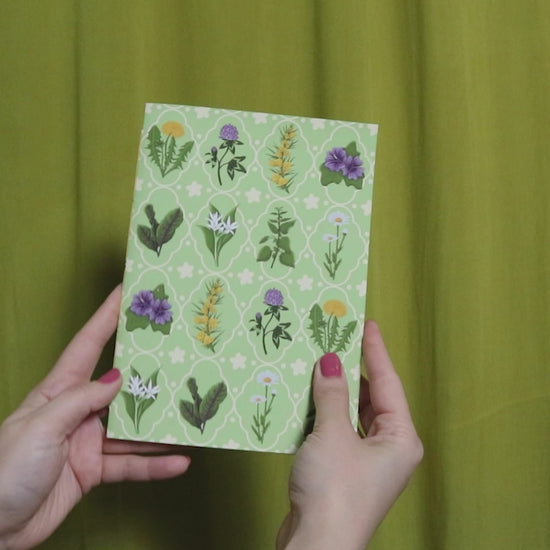 Video of two hands holding and flipping through a green sketchbook with floral pattern against a green curtain background