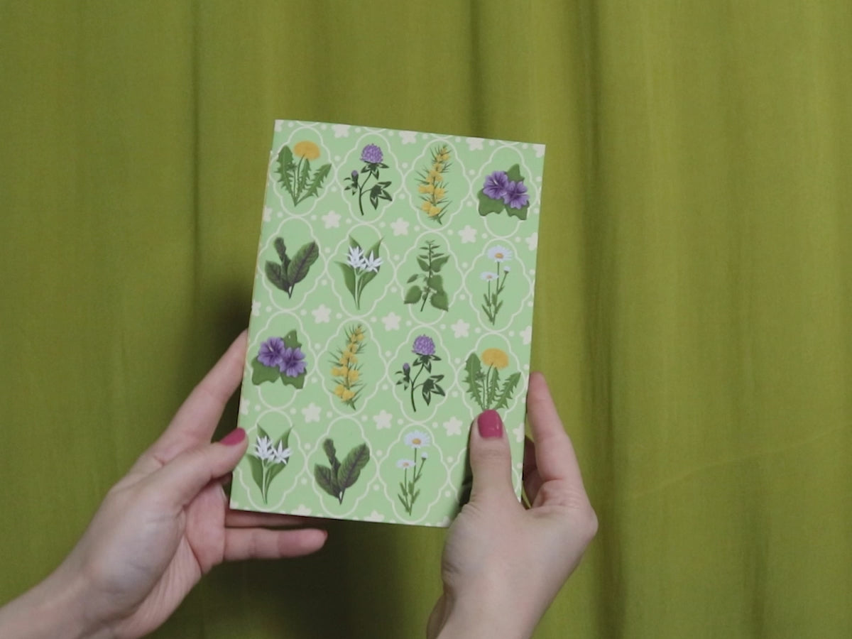 Video of two hands holding and flipping through a green sketchbook with floral pattern against a green curtain background