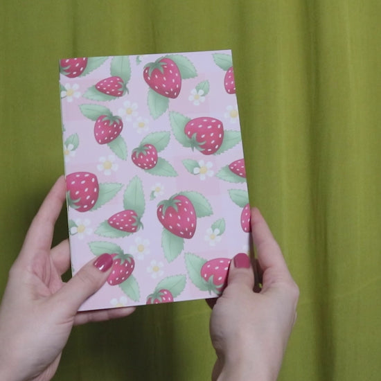 Video of two hands holding and flipping through a sketchbook with strawberry pattern against a green curtain background
