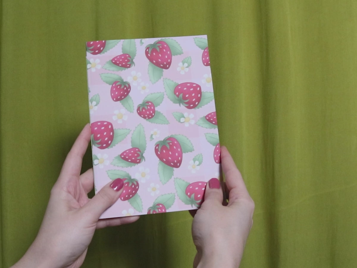 Video of two hands holding and flipping through a sketchbook with strawberry pattern against a green curtain background
