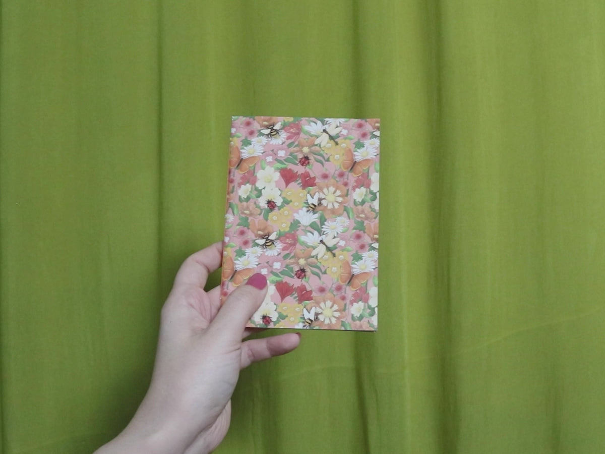 Video of two hands holding and flipping through a small sketchbook with floral pattern against a green curtain background