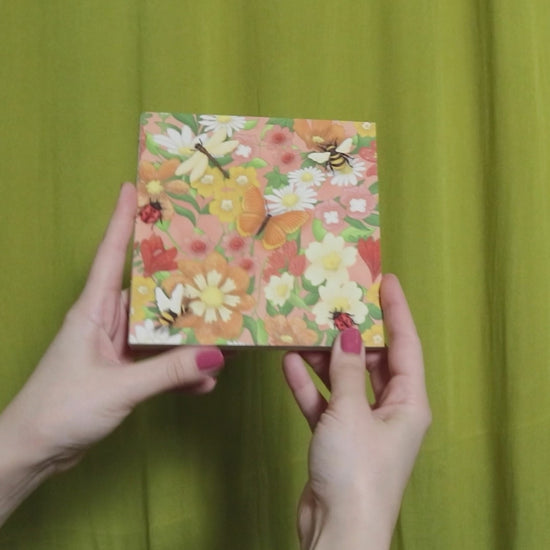 Video of two hands holding and flipping through a square sketchbook with floral pattern against a green curtain background