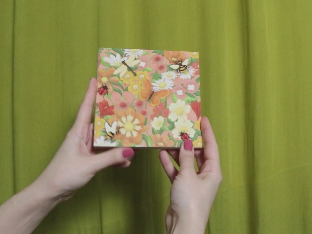 Video of two hands holding and flipping through a square sketchbook with floral pattern against a green curtain background