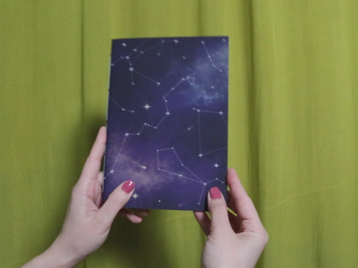 Video of two hands holding and flipping through a sketchbook with cosmic pattern against a green curtain background