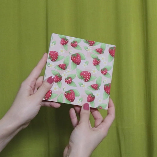 Video of two hands holding and flipping through a square sketchbook with strawberry pattern against a green curtain background
