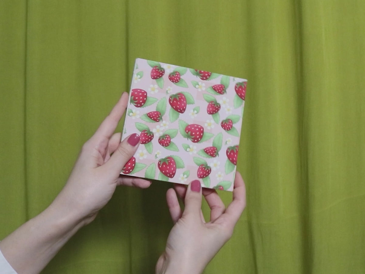 Video of two hands holding and flipping through a square sketchbook with strawberry pattern against a green curtain background