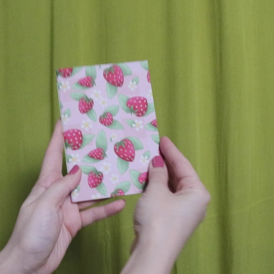 Video of two hands holding and flipping through a small sketchbook with strawberry pattern against a green curtain background