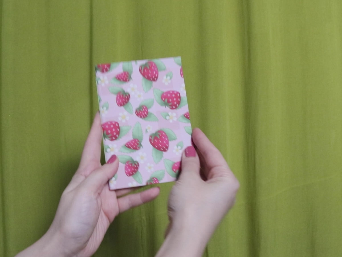 Video of two hands holding and flipping through a small sketchbook with strawberry pattern against a green curtain background