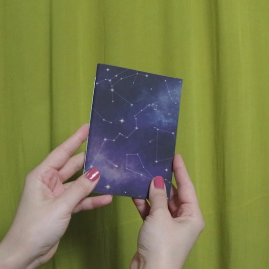 Video of two hands holding and flipping through a small sketchbook with cosmic pattern against a green curtain background
