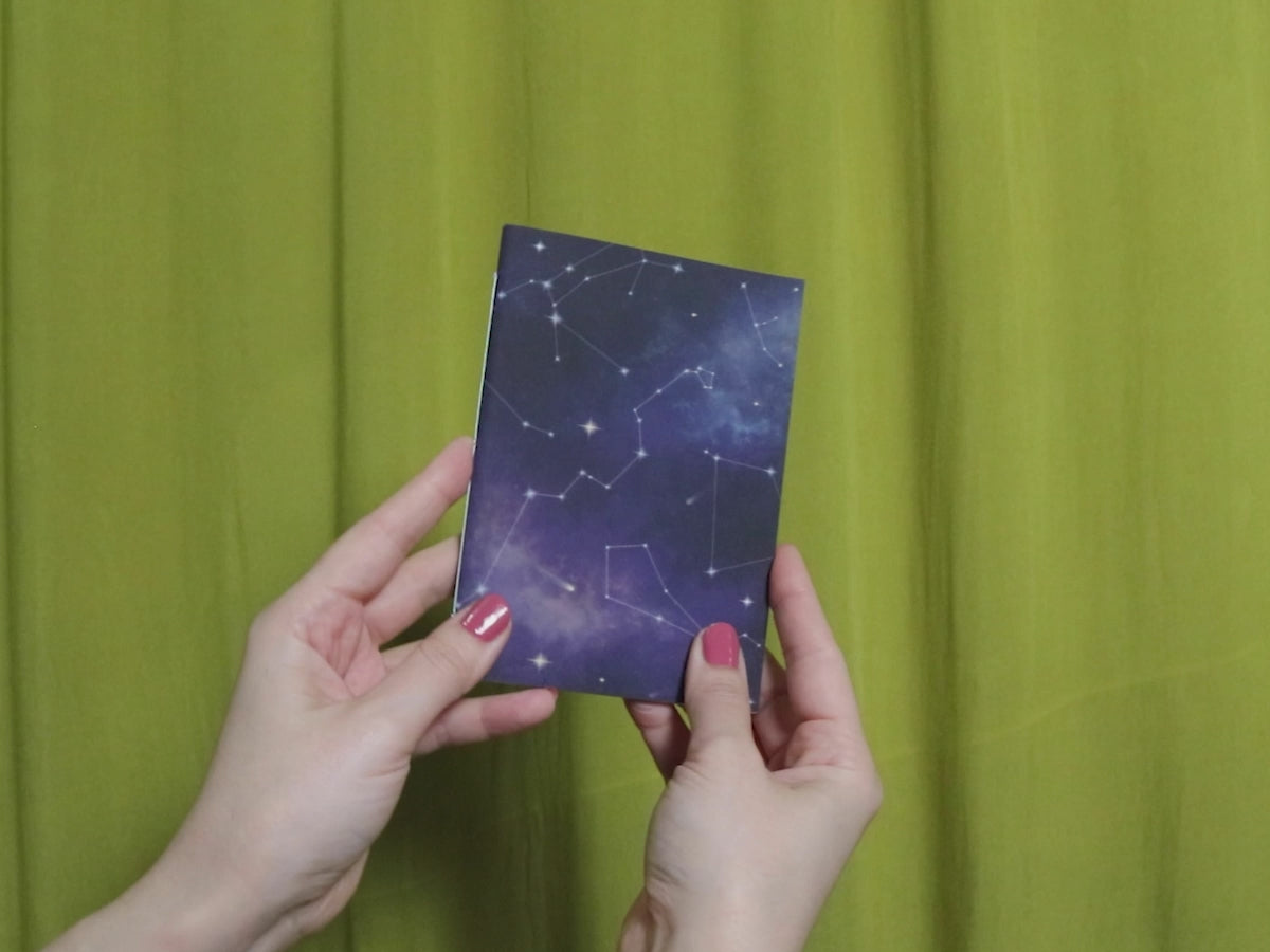 Video of two hands holding and flipping through a small sketchbook with cosmic pattern against a green curtain background