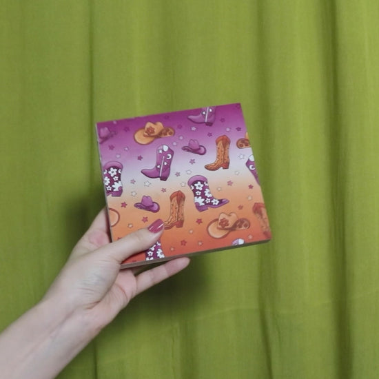 Video of two hands holding and flipping through a square sketchbook with cowgirl pattern against a green curtain background