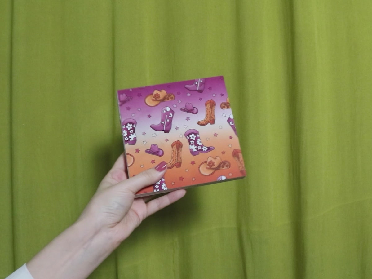 Video of two hands holding and flipping through a square sketchbook with cowgirl pattern against a green curtain background