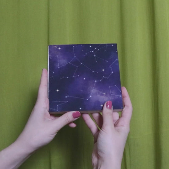 Video of two hands holding and flipping through a square sketchbook with cosmic pattern against a green curtain background