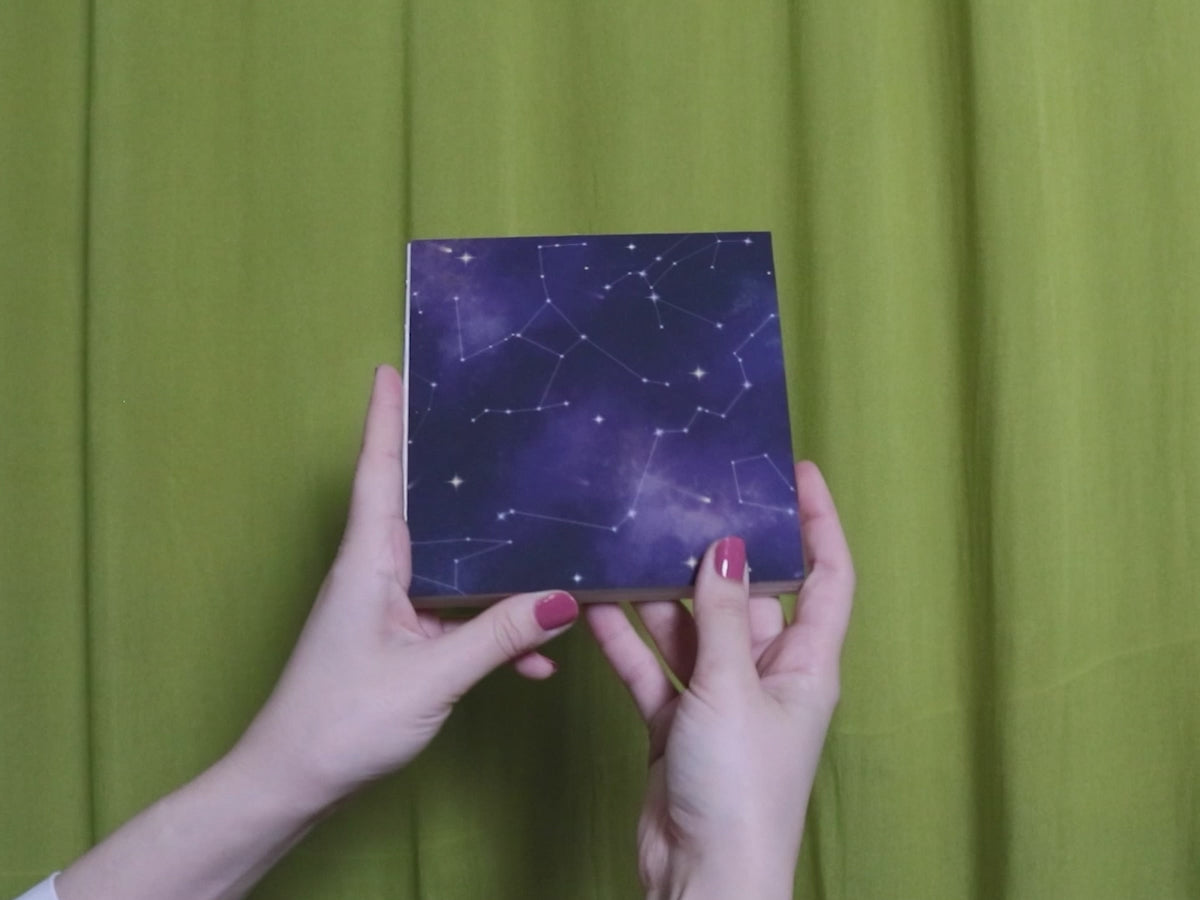 Video of two hands holding and flipping through a square sketchbook with cosmic pattern against a green curtain background
