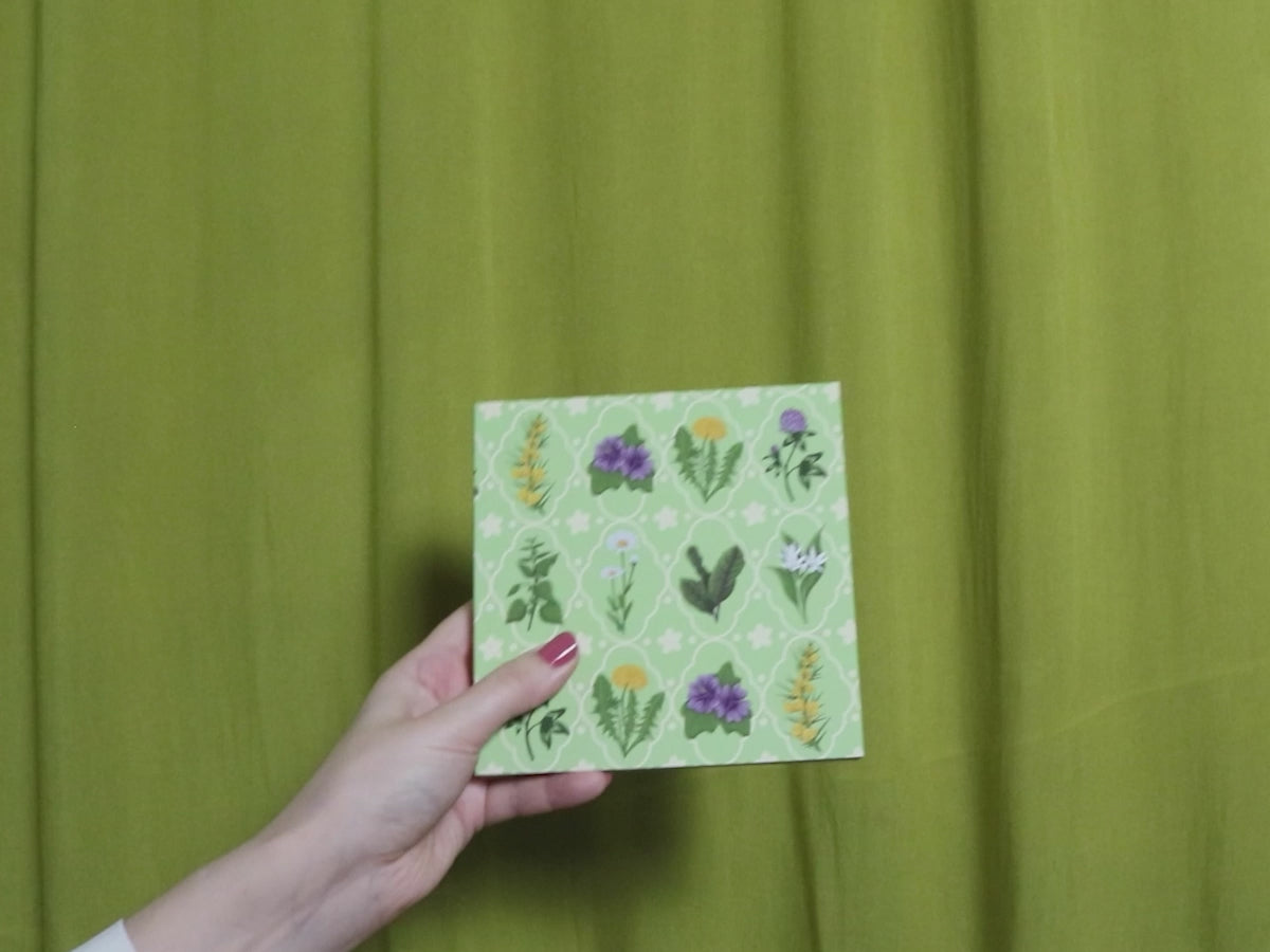 Video of two hands holding and flipping through a square sketchbook with green floral pattern against a green curtain background
