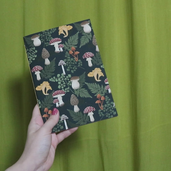 Video of two hands holding and flipping through a sketchbook with mushroom pattern against a green curtain background