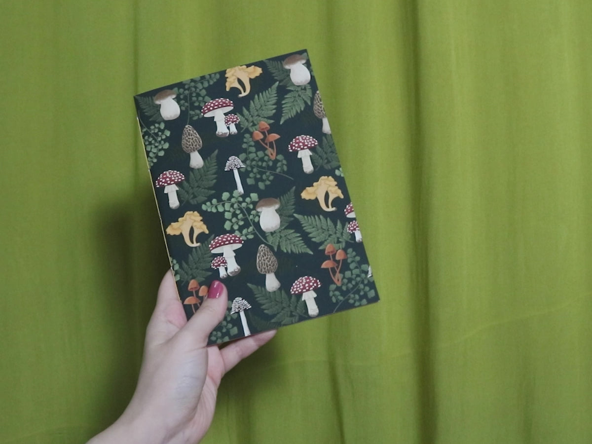 Video of two hands holding and flipping through a sketchbook with mushroom pattern against a green curtain background